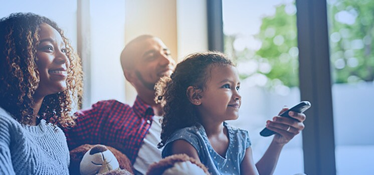 Mother and daughter sitting on couch smiling and using a remote while watching TV.